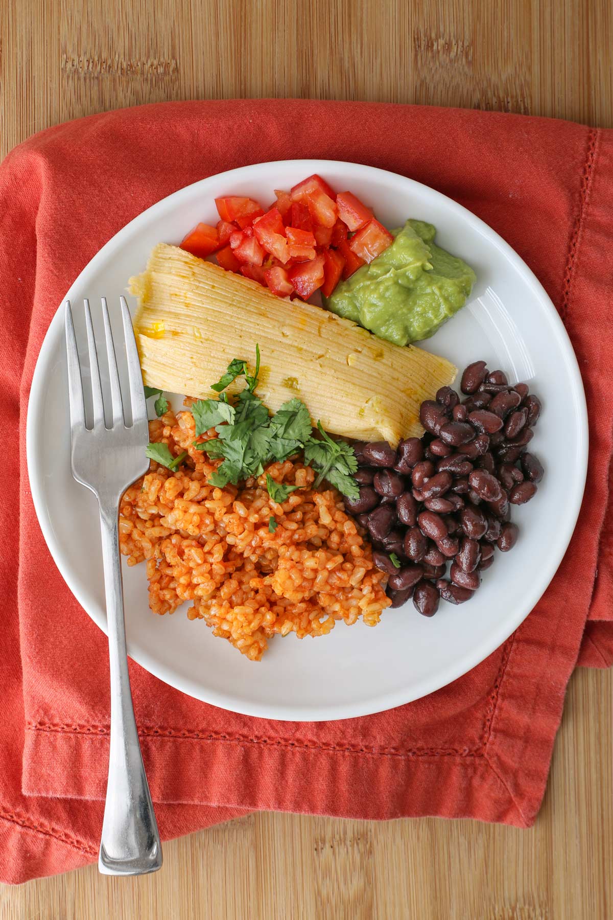 dinner plate with a cheese tamale, rice, beans, and condiments, on an orange napkin.