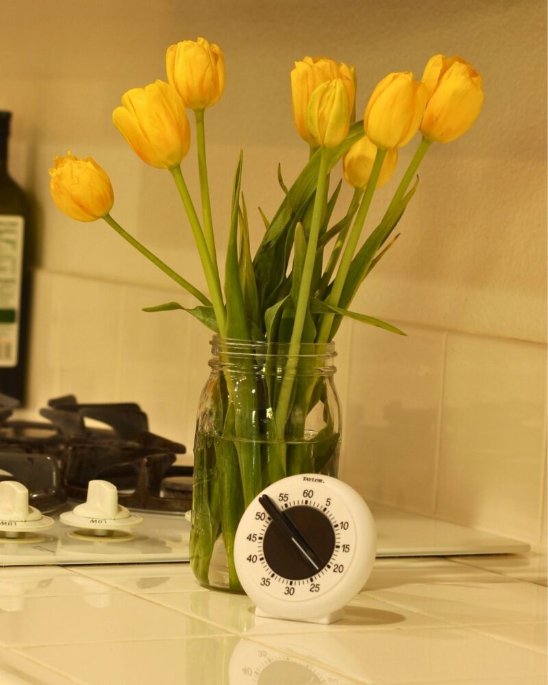 jar of yellow tulips and a white kitchen timer on white tile counter near stovetop.