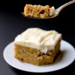 close up of fork of spiced zucchini cake with cream cheese frosting above a square of cake on a white plate.