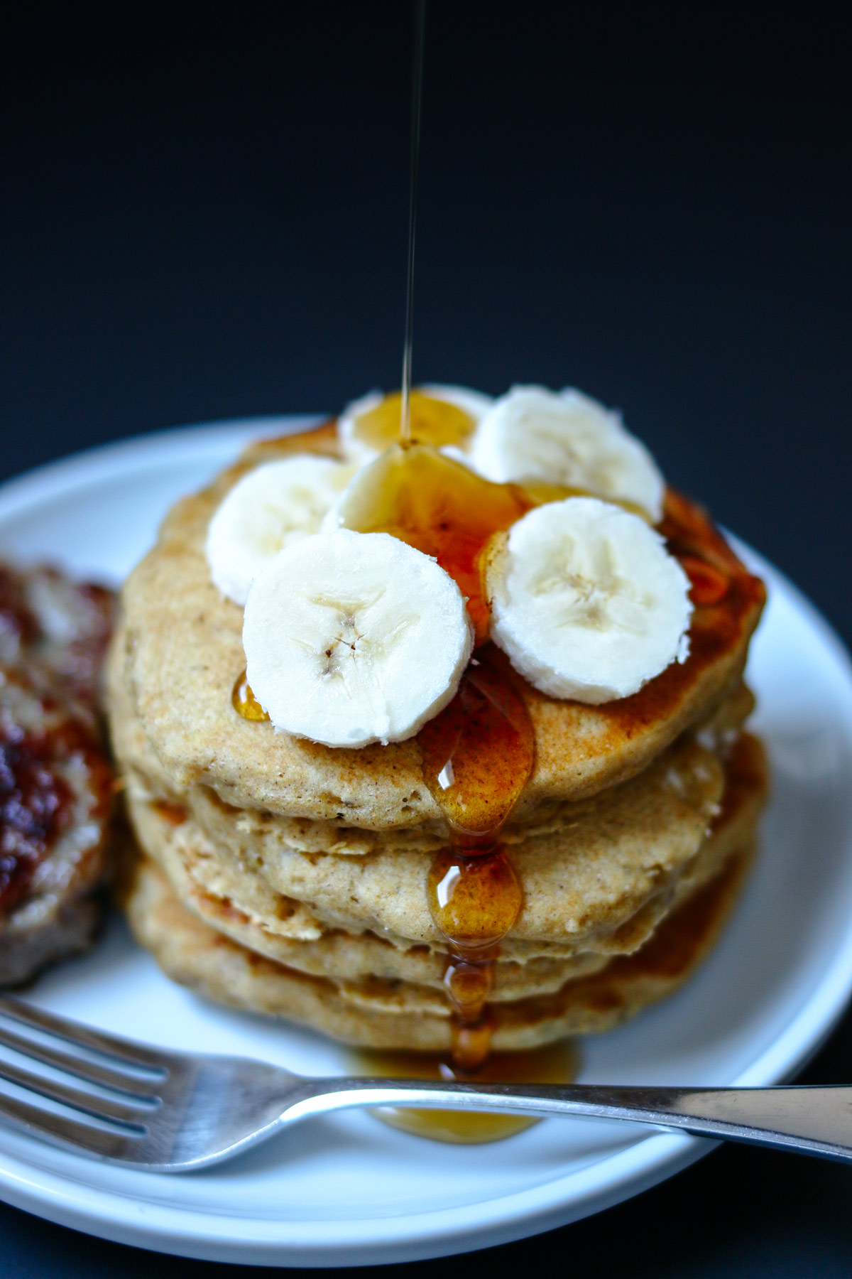 shot of maple syrup pouring onto banana slices atop whole wheat pancakes on a plate with sausage patties and a fork.