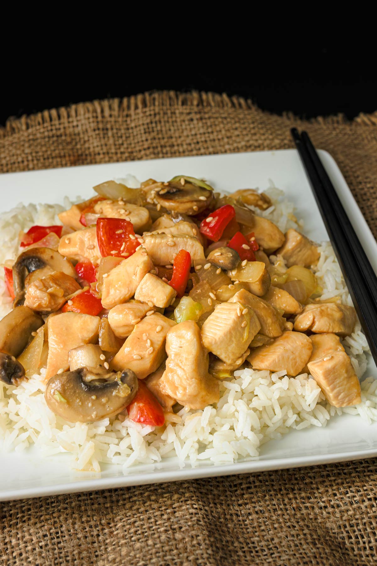 chicken stir fry on a bed of rice on a white square plate with black chopsticks.