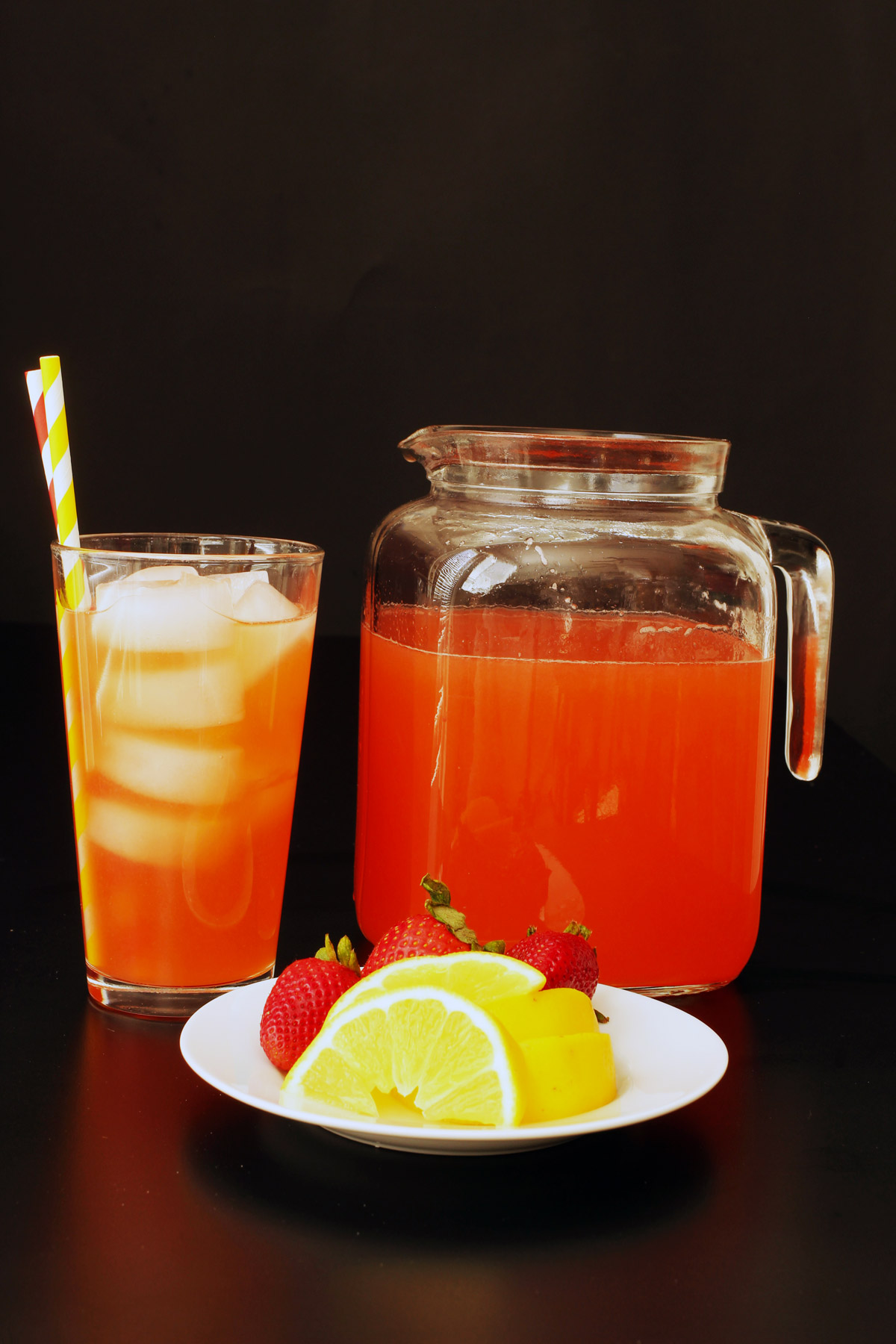 sideview of glass of lemonade, full pitcher, and plate of strawberries and lemon wedges