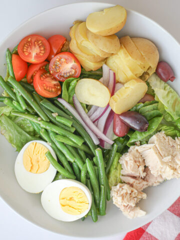 salad nicoise in a white bowl next to a red check cloth with a fork.