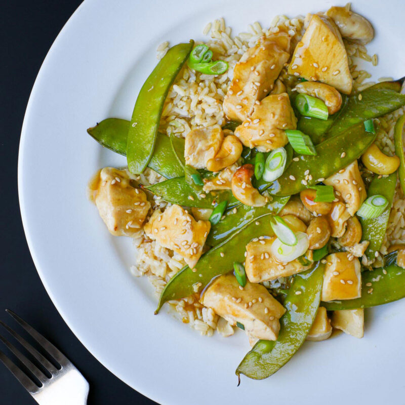 overhead shot of cashew chicken on a white dinner plate with a fork nearby on the black table.