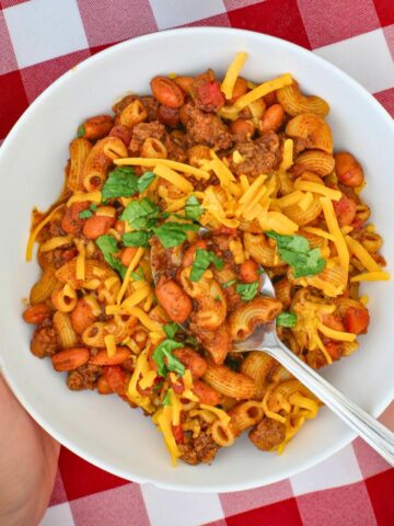 hands holding a white bowl of chili mac with a spoon and toppings.