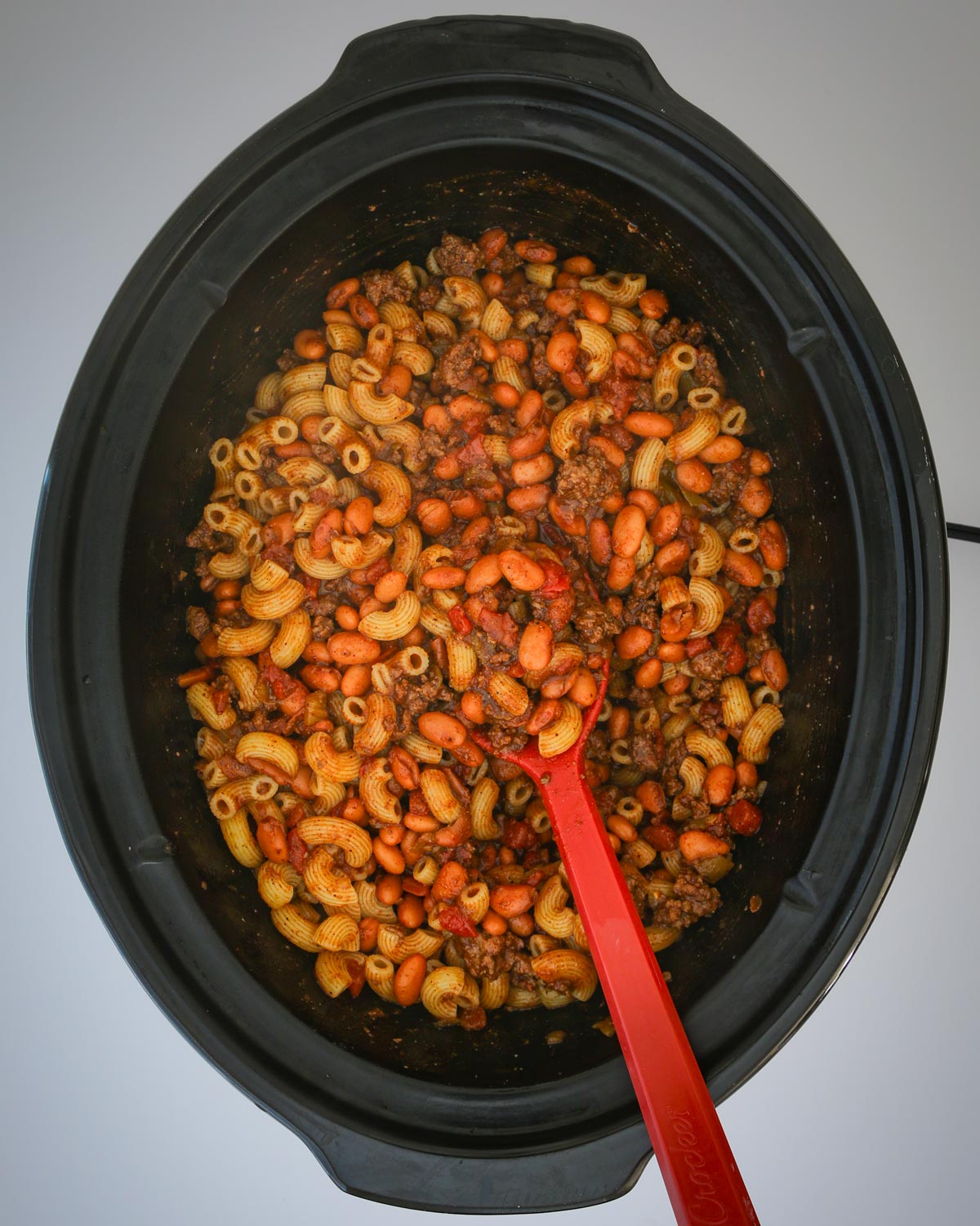 chili mac in the slow cooker with a red spoon.