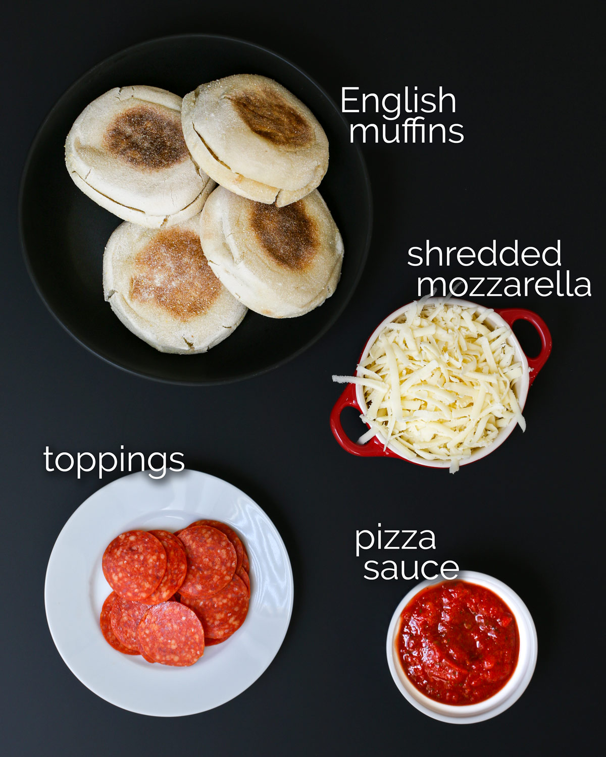 ingredients for English muffin pizzas laid out on a black table.