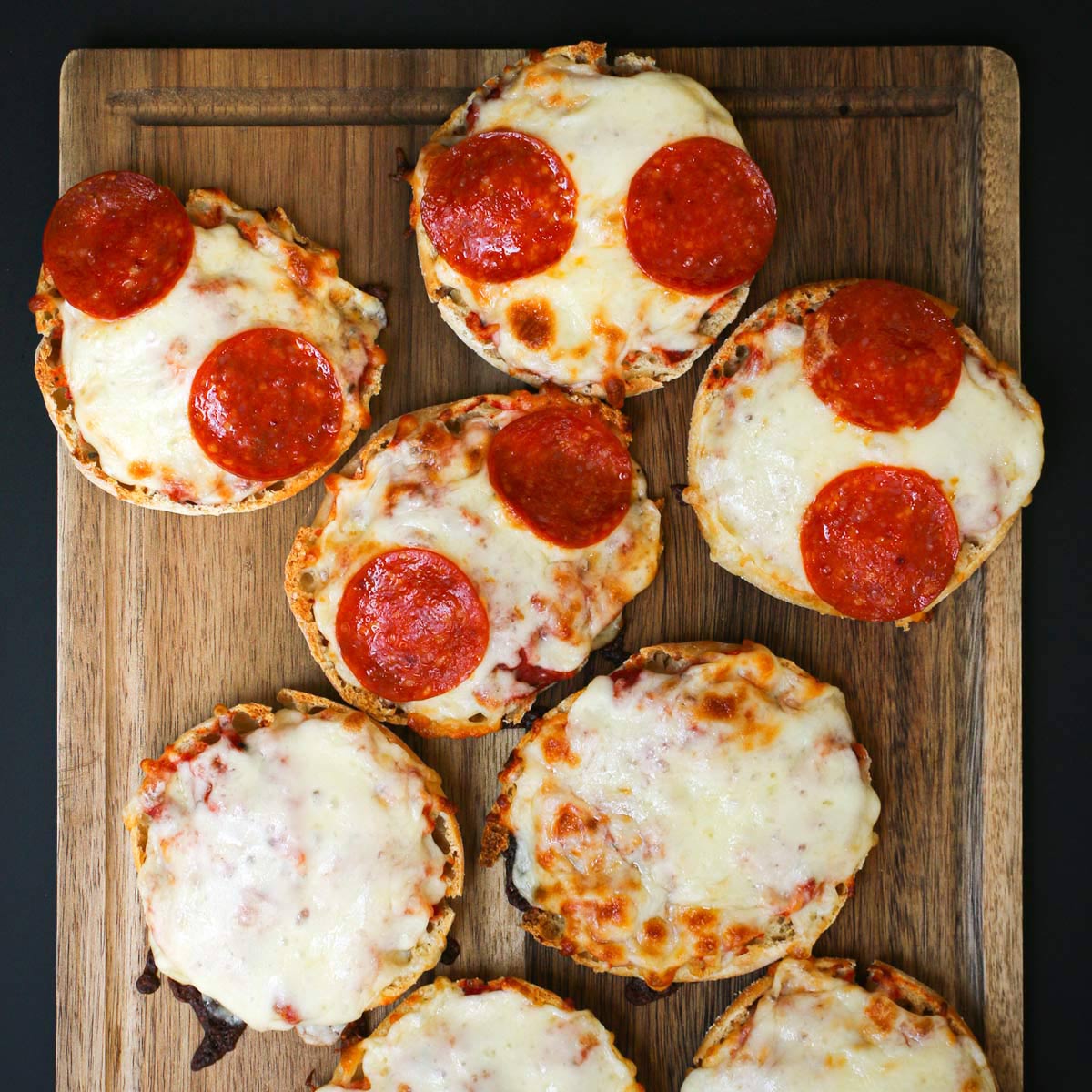 overhead shot of english muffin pizzas, some pepperoni some cheese, on a wooden board.