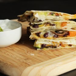 A quesadilla sitting on top of a wooden cutting board, with dish of sour cream