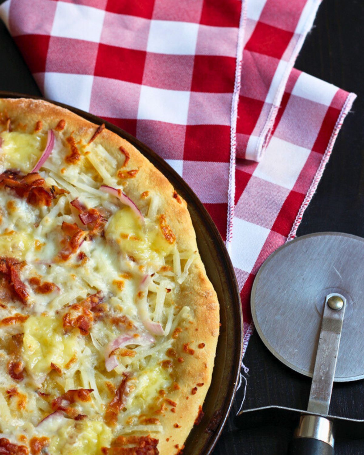 overhead shot of tartiflette pizza, red checked cloth, and a pizza wheel.