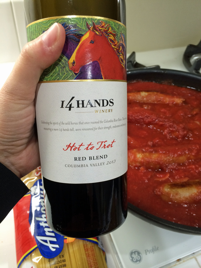A person holding a bottle of wine next to pan of ragu