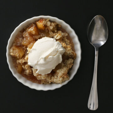 close up of apple crisp with scoop of whipped cream in a ramekin with a spoon nearby.