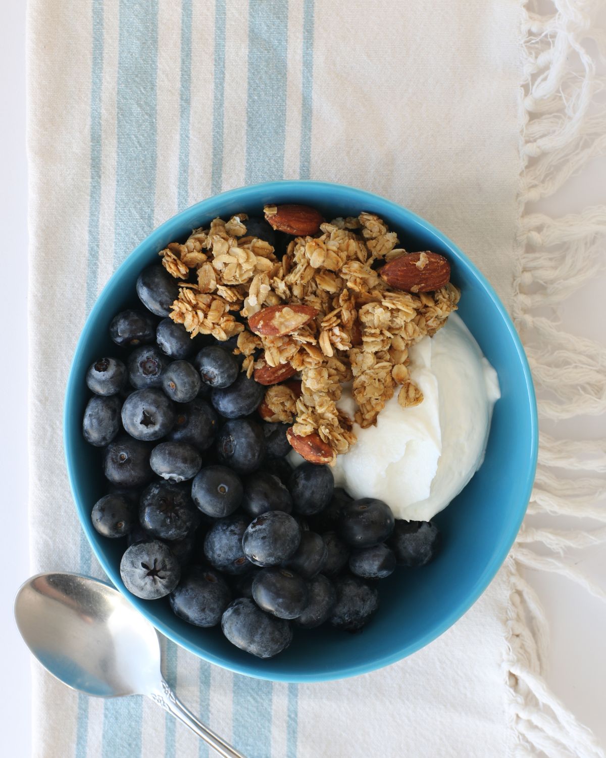 teal bowl of blueberries, yogurt, and granola on a blue striped cloth with a spoon on the side.
