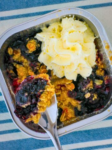 overhead shot of blueberry crumble with a spoon scooping out a bite, whipped cream on top.