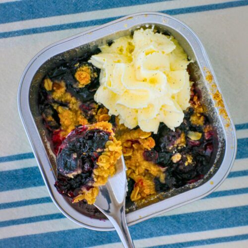 overhead shot of blueberry crumble with a spoon scooping out a bite, whipped cream on top.