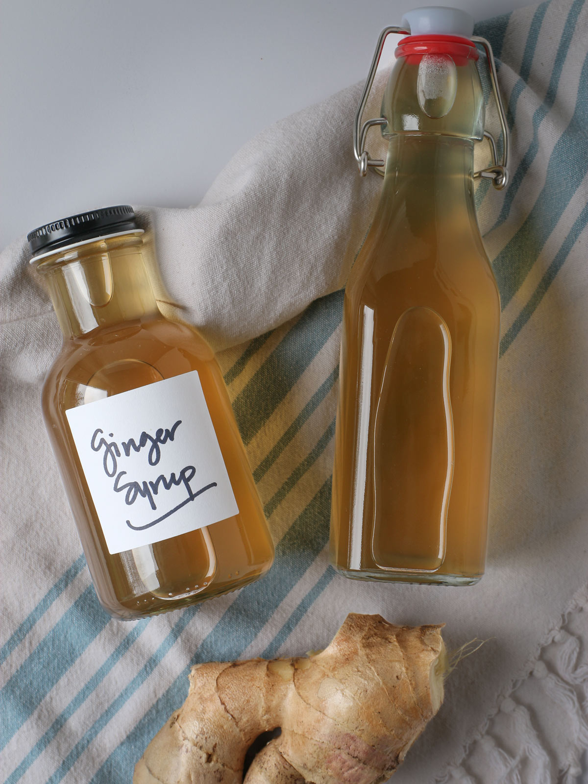 two bottles of ginger syrup with a large chunk of ginger root, laid out on a blue and white striped towel.