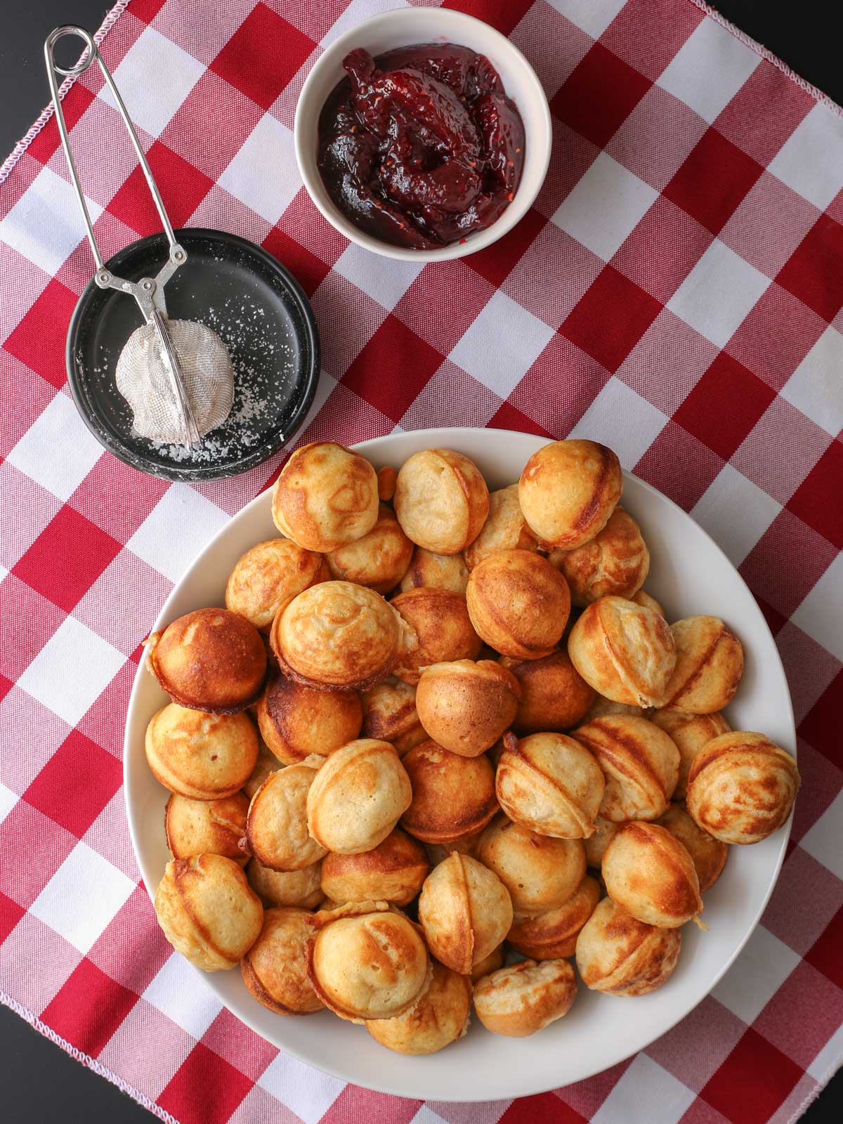 large platter full of aebleskiver with toppings in dishes nearby on checked cloth table.