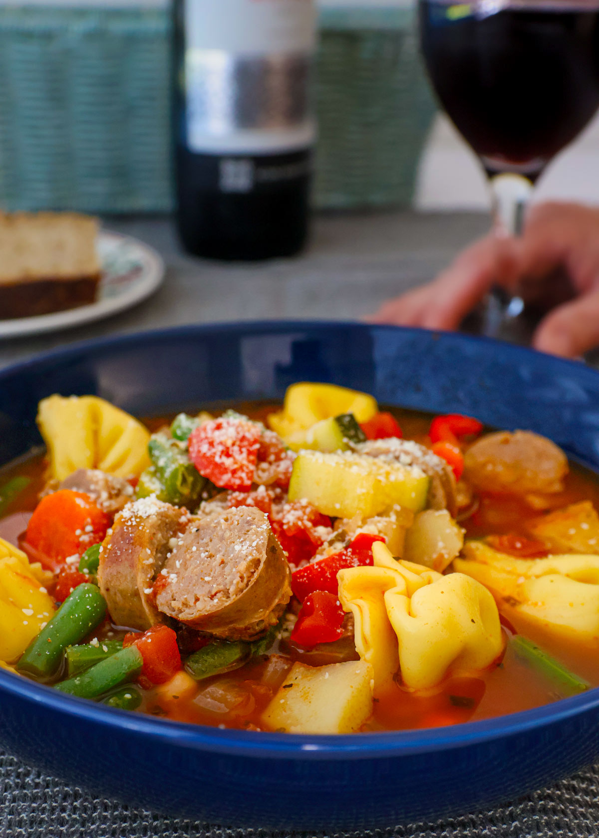 side view of a bowl of minestrone with a hand holding a glass of red wine in the background.