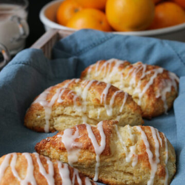 orange scones in a tray lined with a blue cloth, in front of a bowl of oranges.