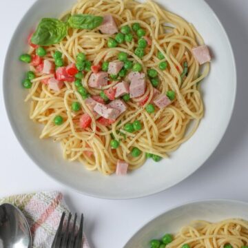 two plates of ham pasta with forks and spoons on table nearby.