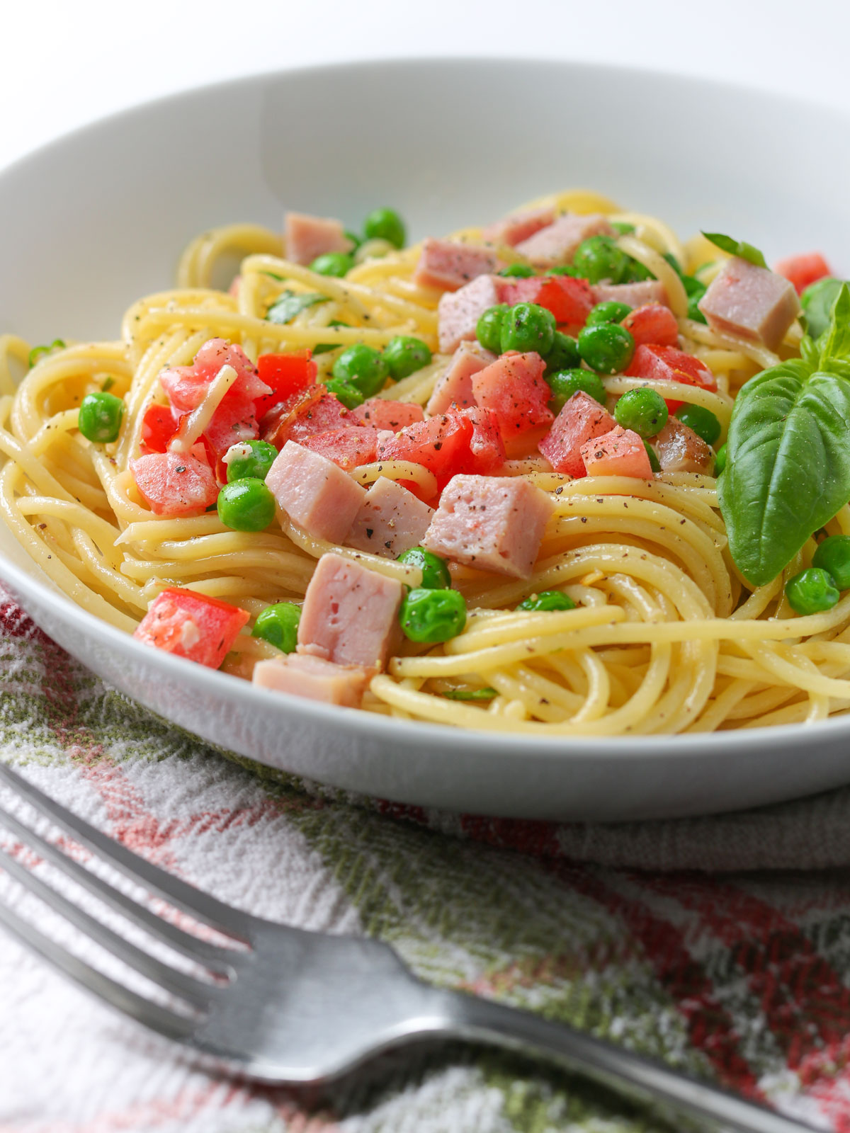 side view of a bowl of ham pasta with peas, with a fork and checked cloth.