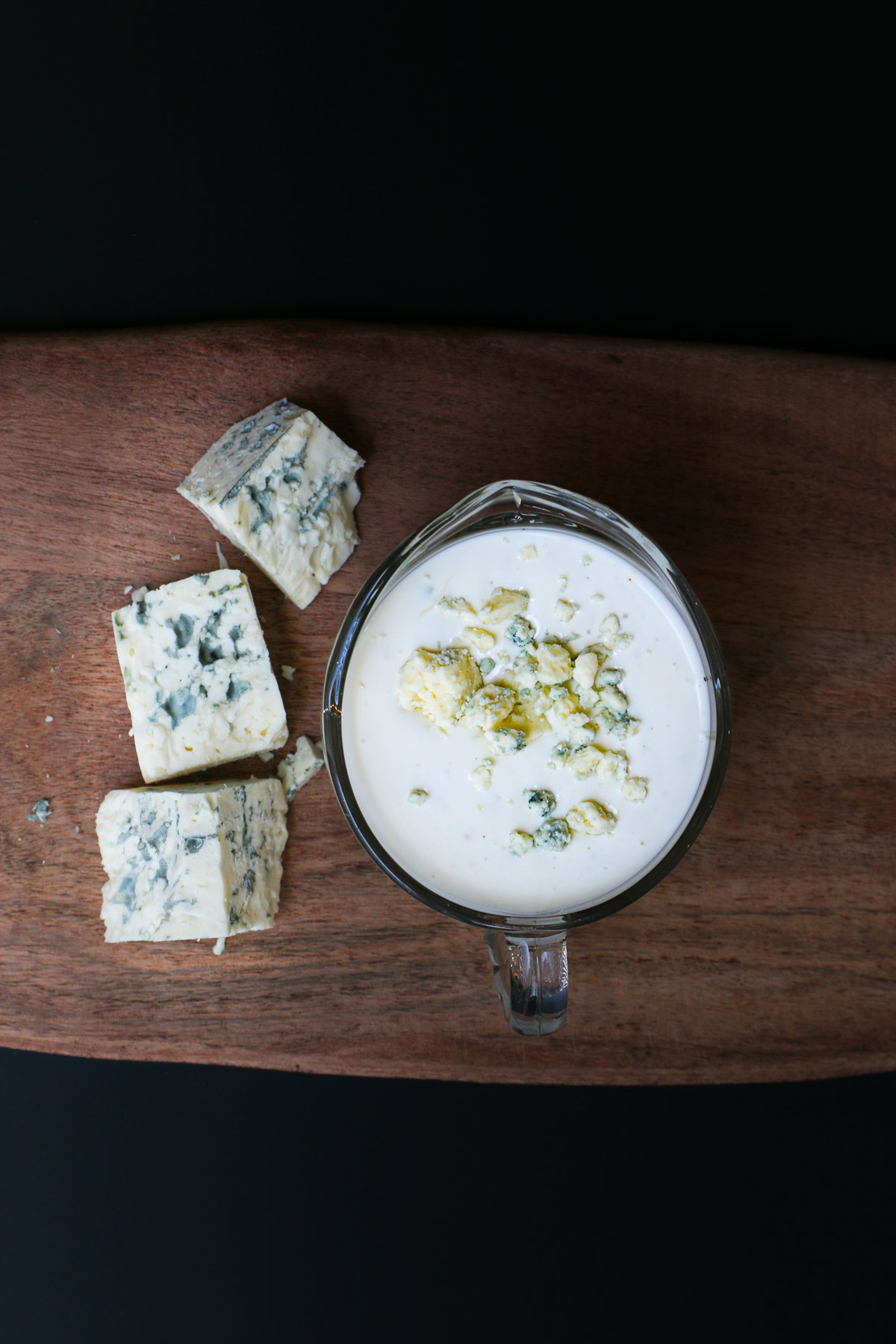 overhead shot of blue cheese dressing in a small pitcher on a wooden board with three chunks of blue cheese nearby.