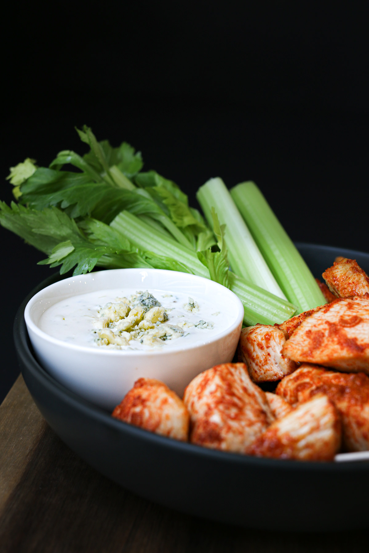 sideview shot of platter of celery and buffalo chicken bites with a small dish of blue cheese dressing.