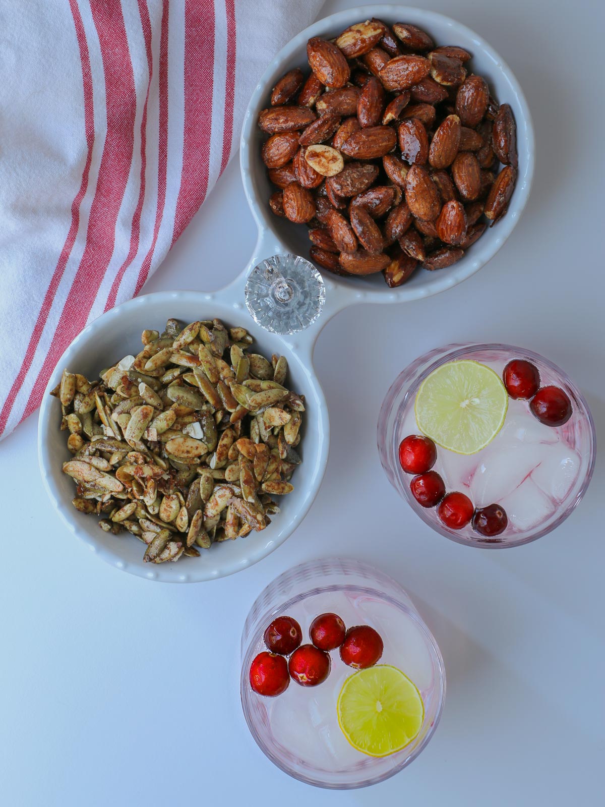 bowls of candied almonds and pipits next to cocktails on white table with cloth.