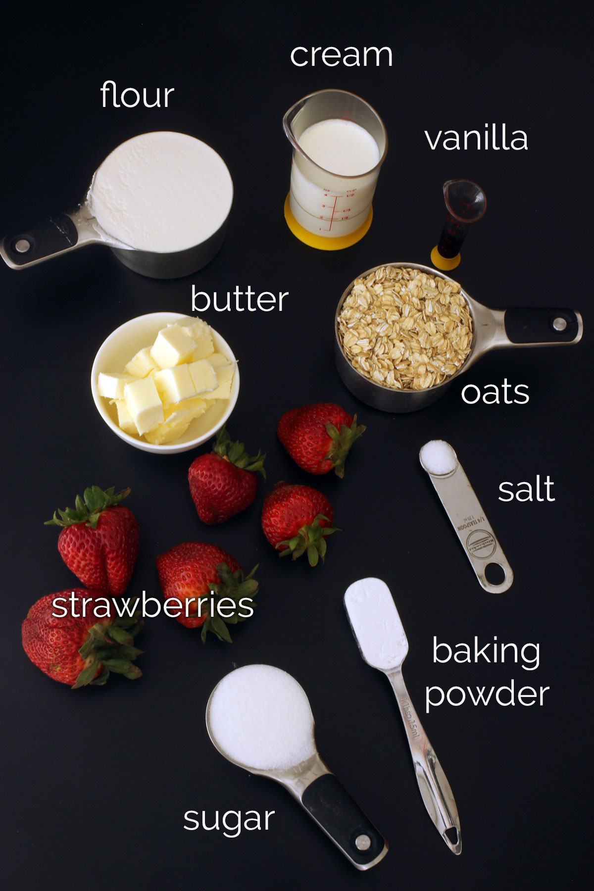 ingredients for strawberry oatcakes in measuring cups laid out on a black table.