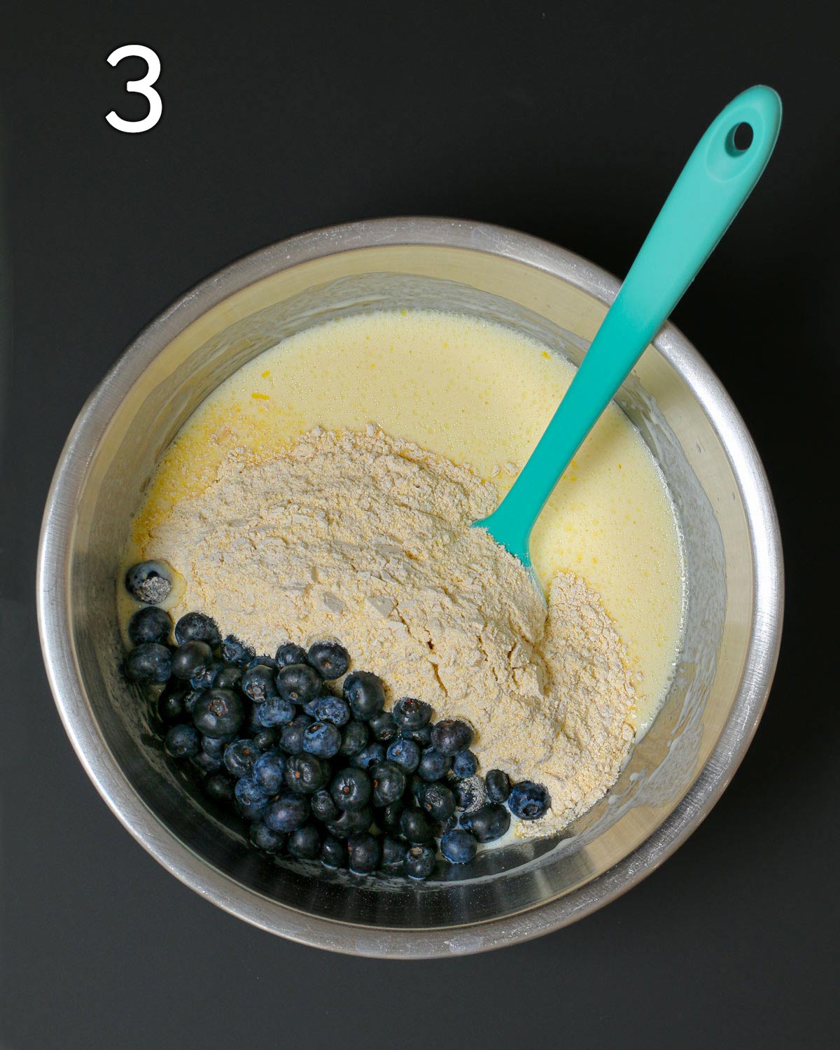 dry added to wet in bowl with blueberries atop; a teal spatula is submerged in it all.
