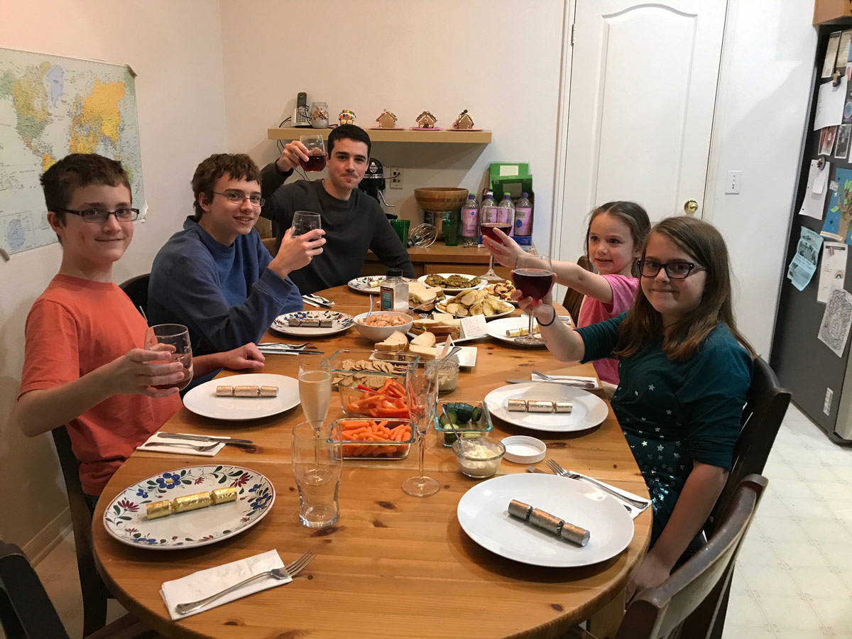 five kids around kitchen table toasting with juice in wine glasses.