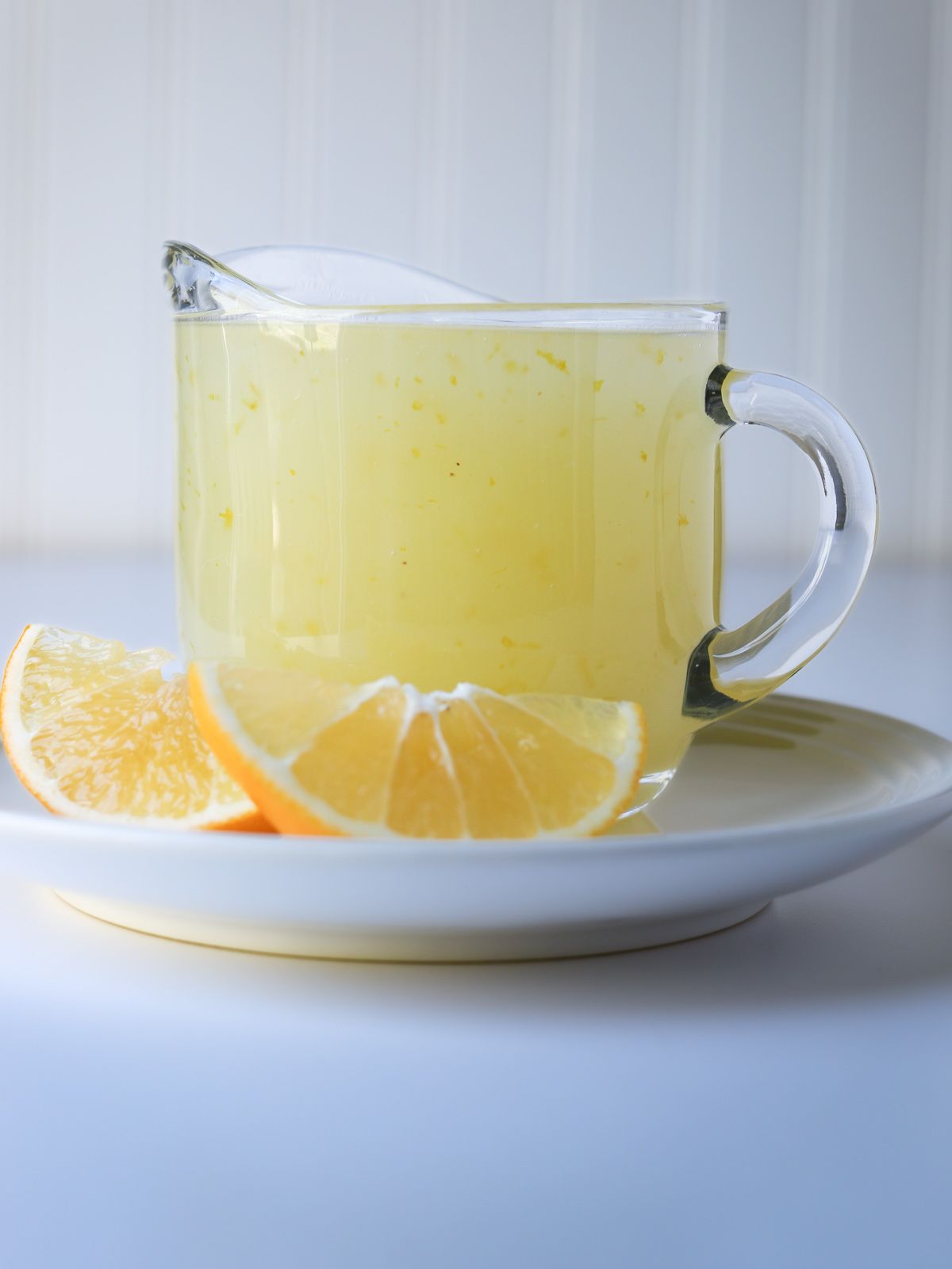 small glass pitcher of lemon sauce on a saucer with lemon wedges.