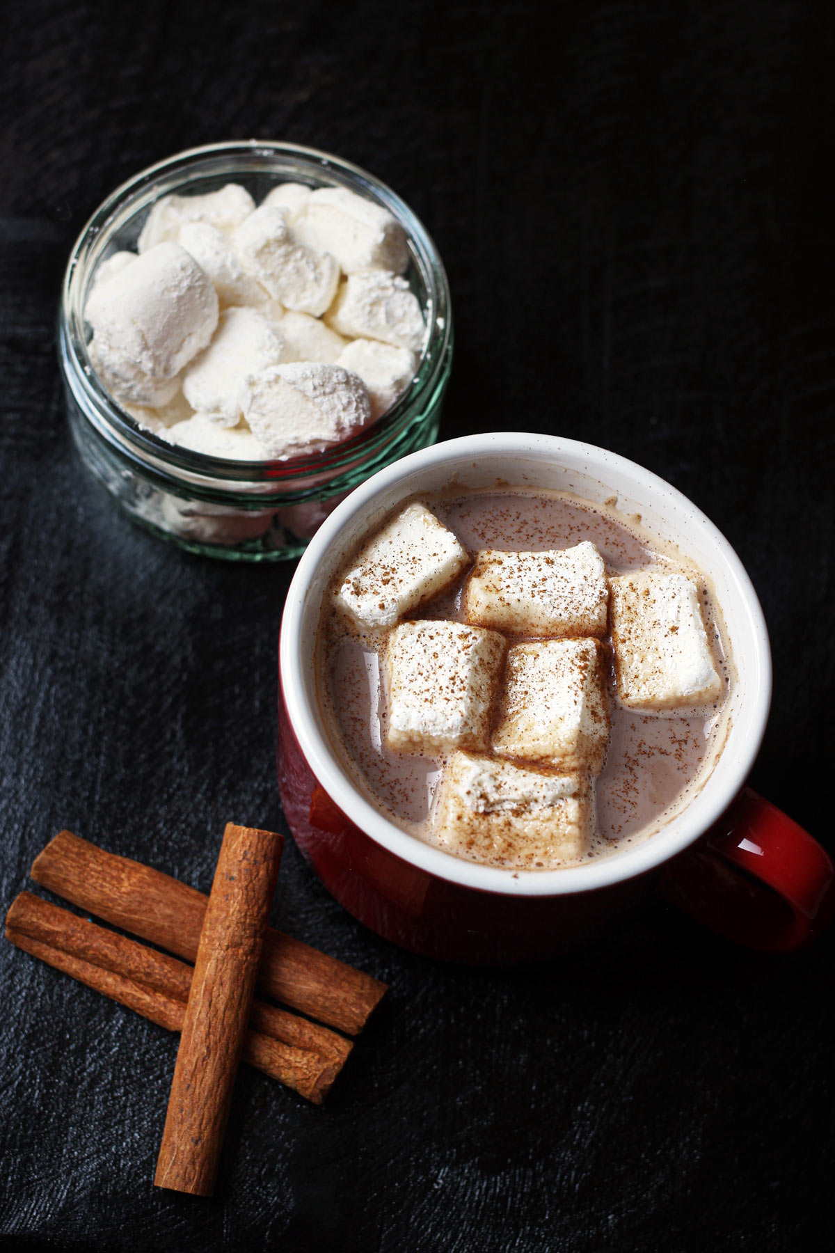 mug of hot cocoa on black table with marshmallows and cinnamon sticks.