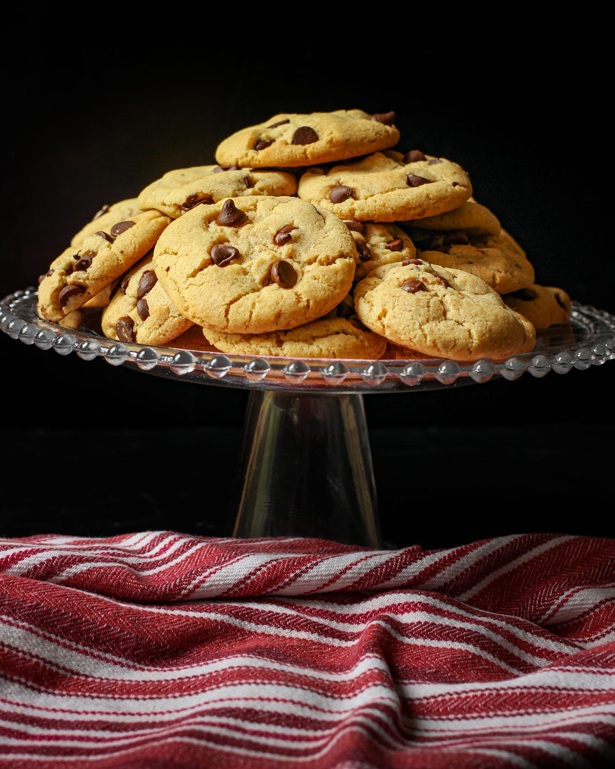 pile of chocolate chip cookies on cake platter with red striped cloth nearby.