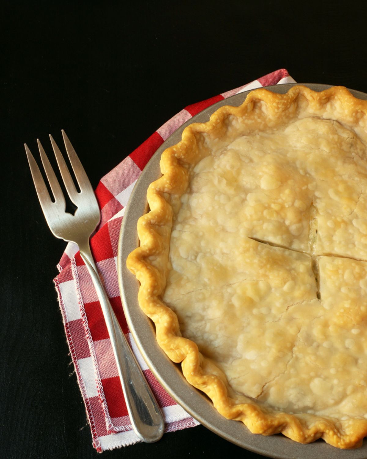 chicken pot pie on red checked cloth with fork on black table.