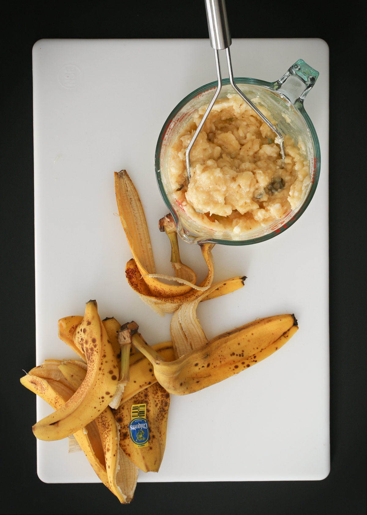 mashing bananas in glass pyrex measure with a potato masher on a white cutting board on a black table.