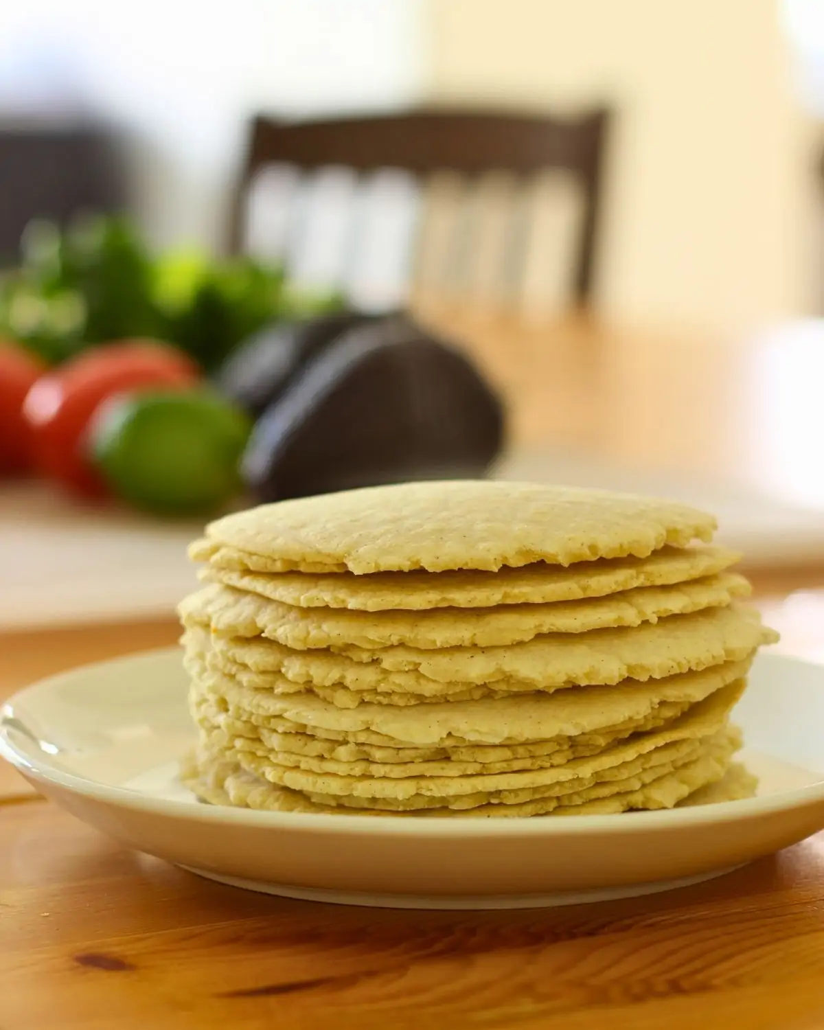 stack of corn tortillas on table with avocado, tomato, and limes in background.