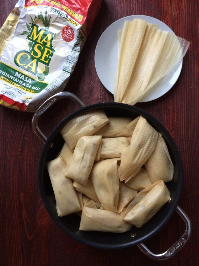 tamales stacked in steaming pot with maseca and corn husks nearby