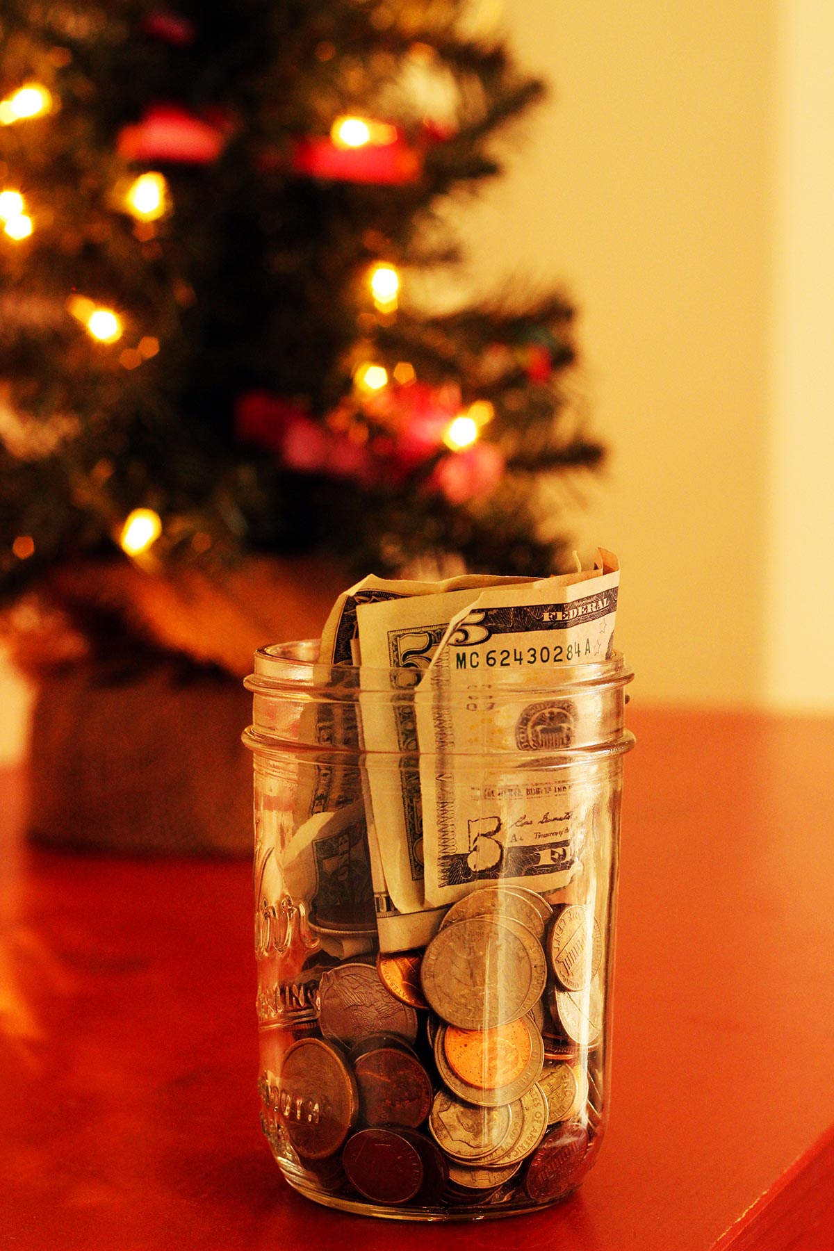 lit mini christmas tree on red table top with a jar of coins and US dollars.