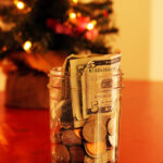 jar of money on table in front of a small lit christmas tree.