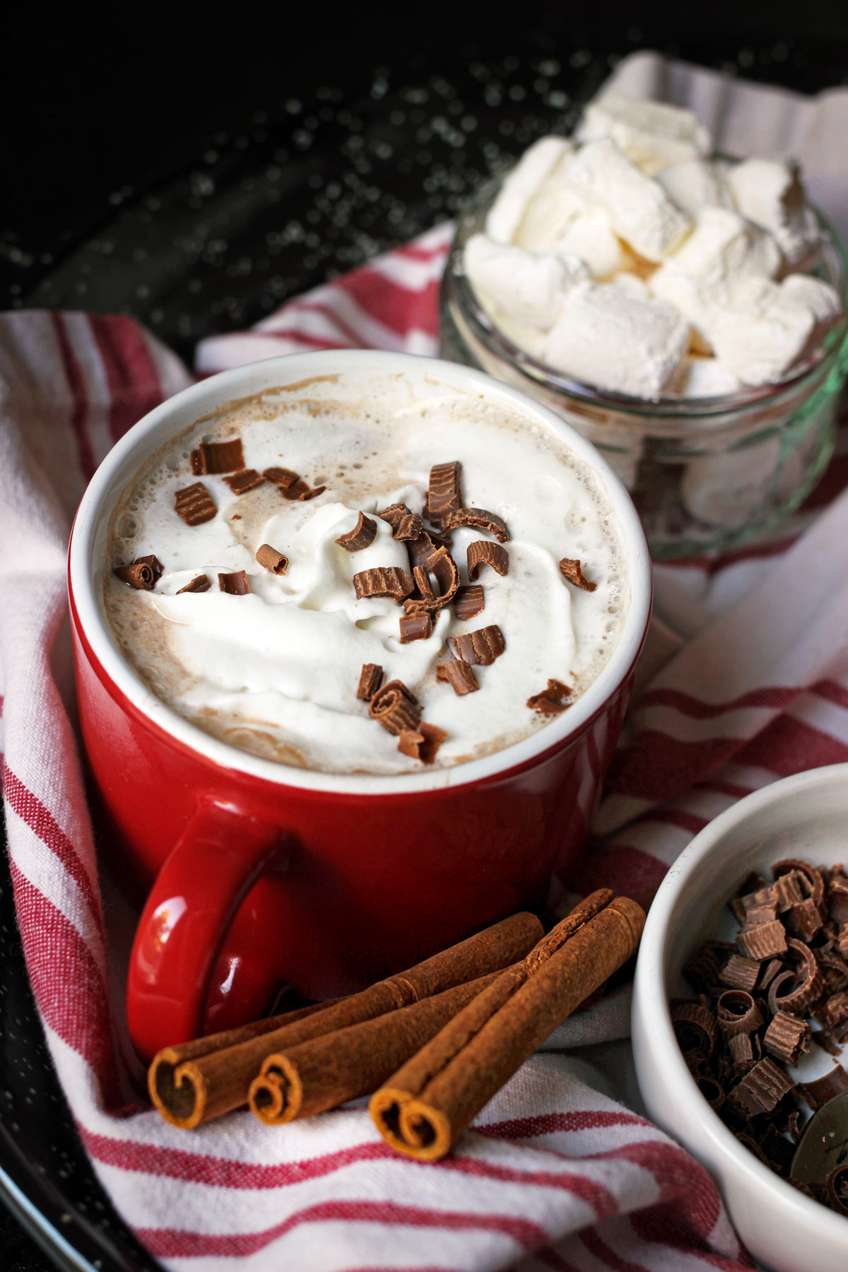 red mug of cocoa topped with whipped cream and chocolate shavings with a dish of homemade marshmallows in the background.
