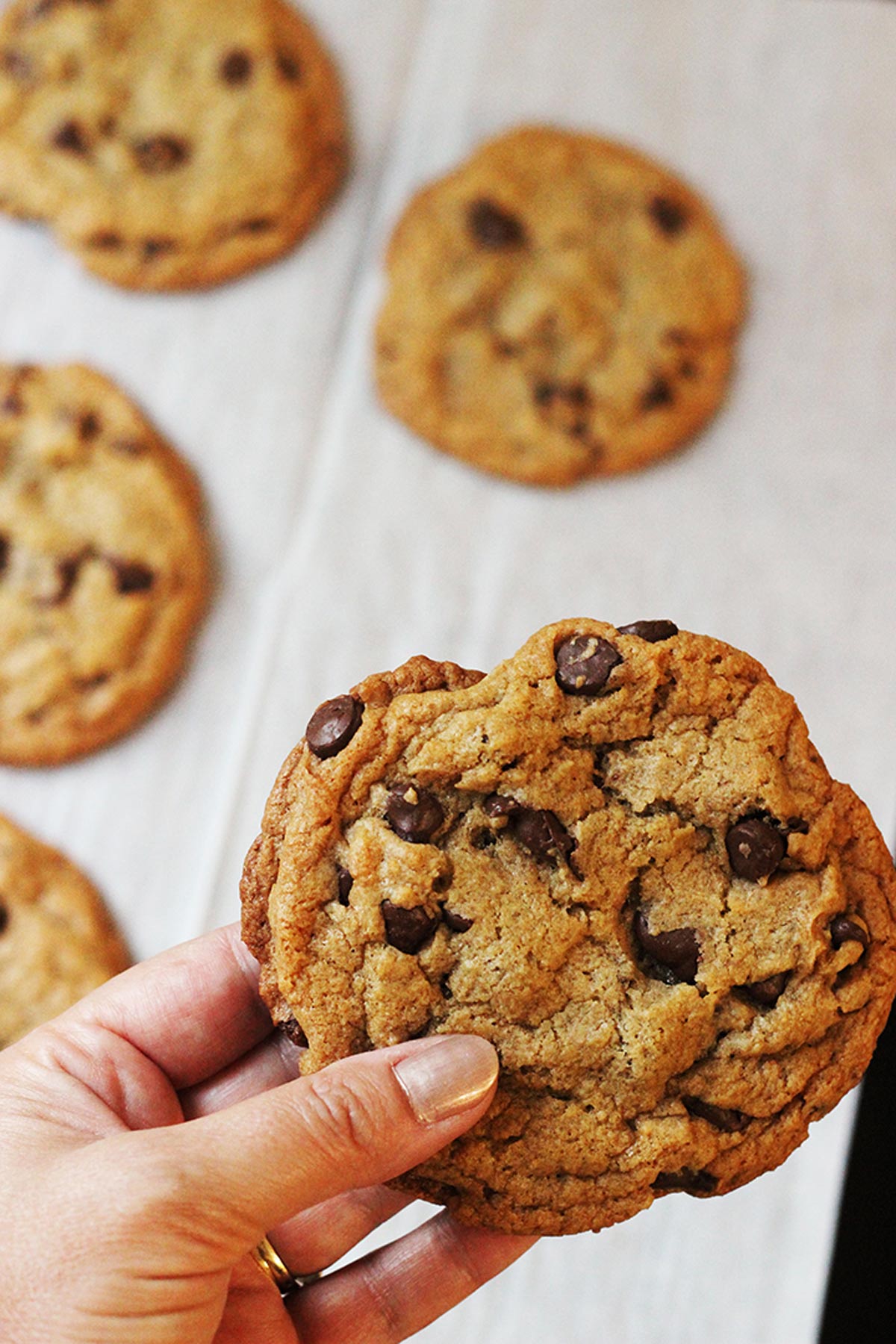 woman holding jump chocolate chip cookie above a pan of other cookies.