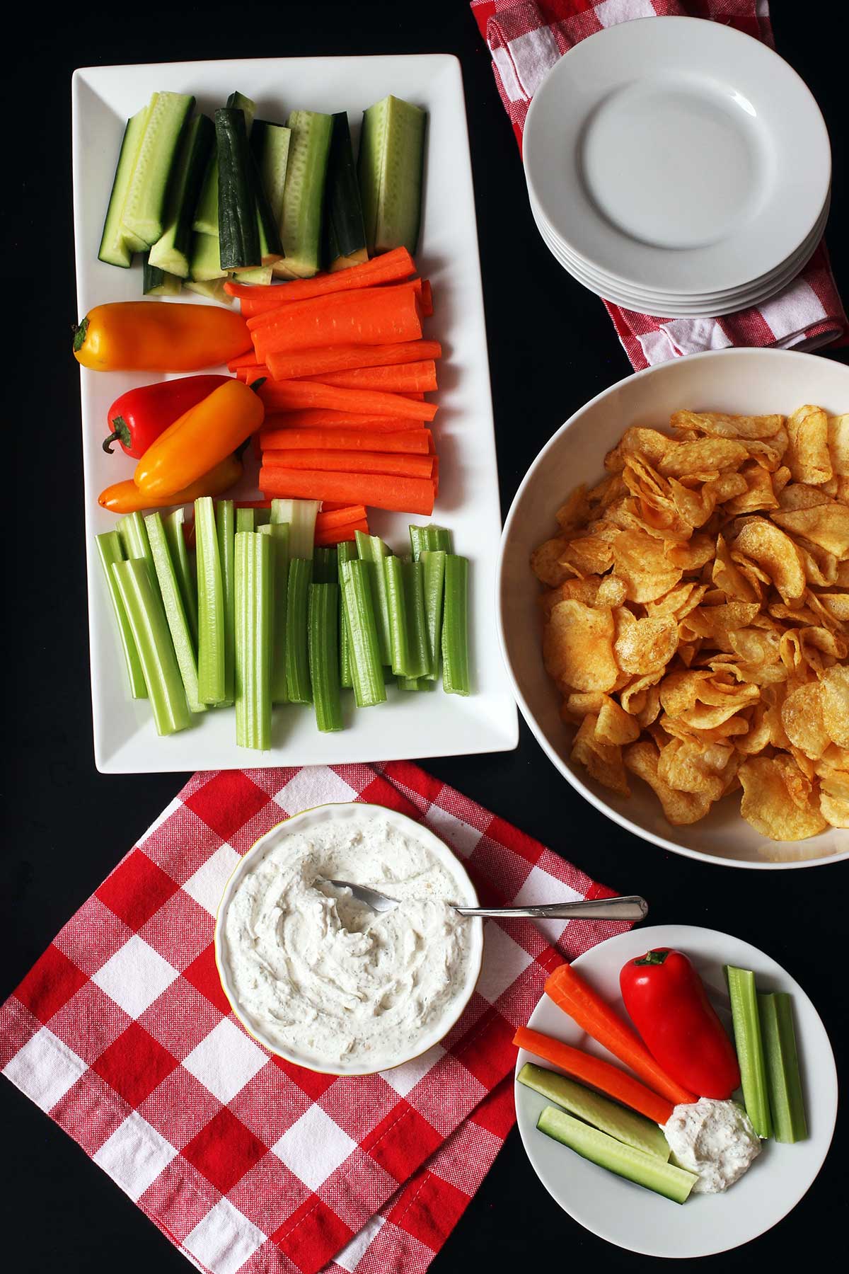 appetizer table set with veggies, chips, and dill dip with a red checked napkin.