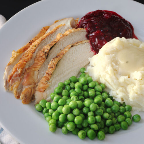 thanksgiving dinner plate featuring slices of turkey, green peas, mashed potatoes and gravy, and cranberry sauce.