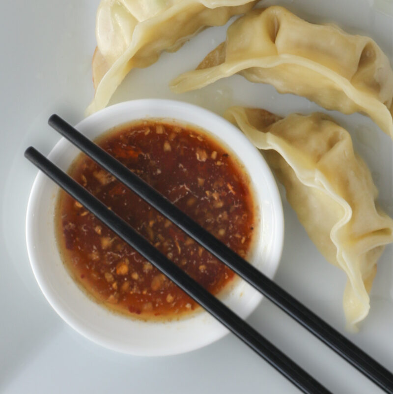 closeup of potsticker sauce in dipping cup with chopsticks and dumplings nearby.