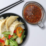 close up of rice bowl with potstickers and a pair of chopsticks, dish of dipping sauce on the side.