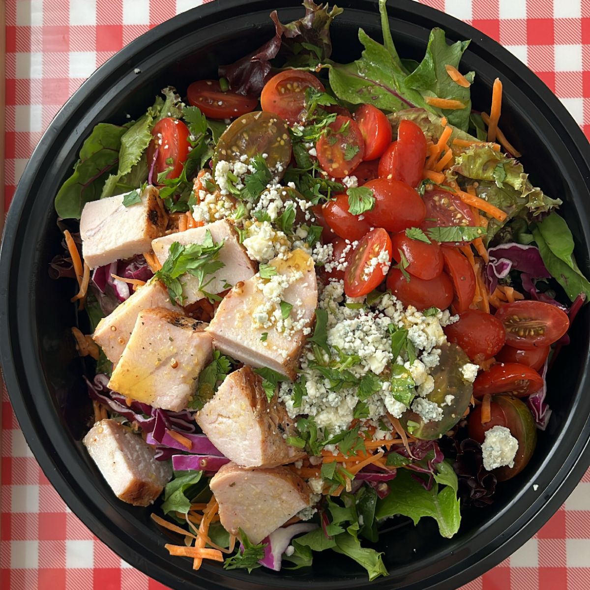 salad with turkey, tomatoes, cheese, and fresh herbs in a black meal prep box on a red checked background.