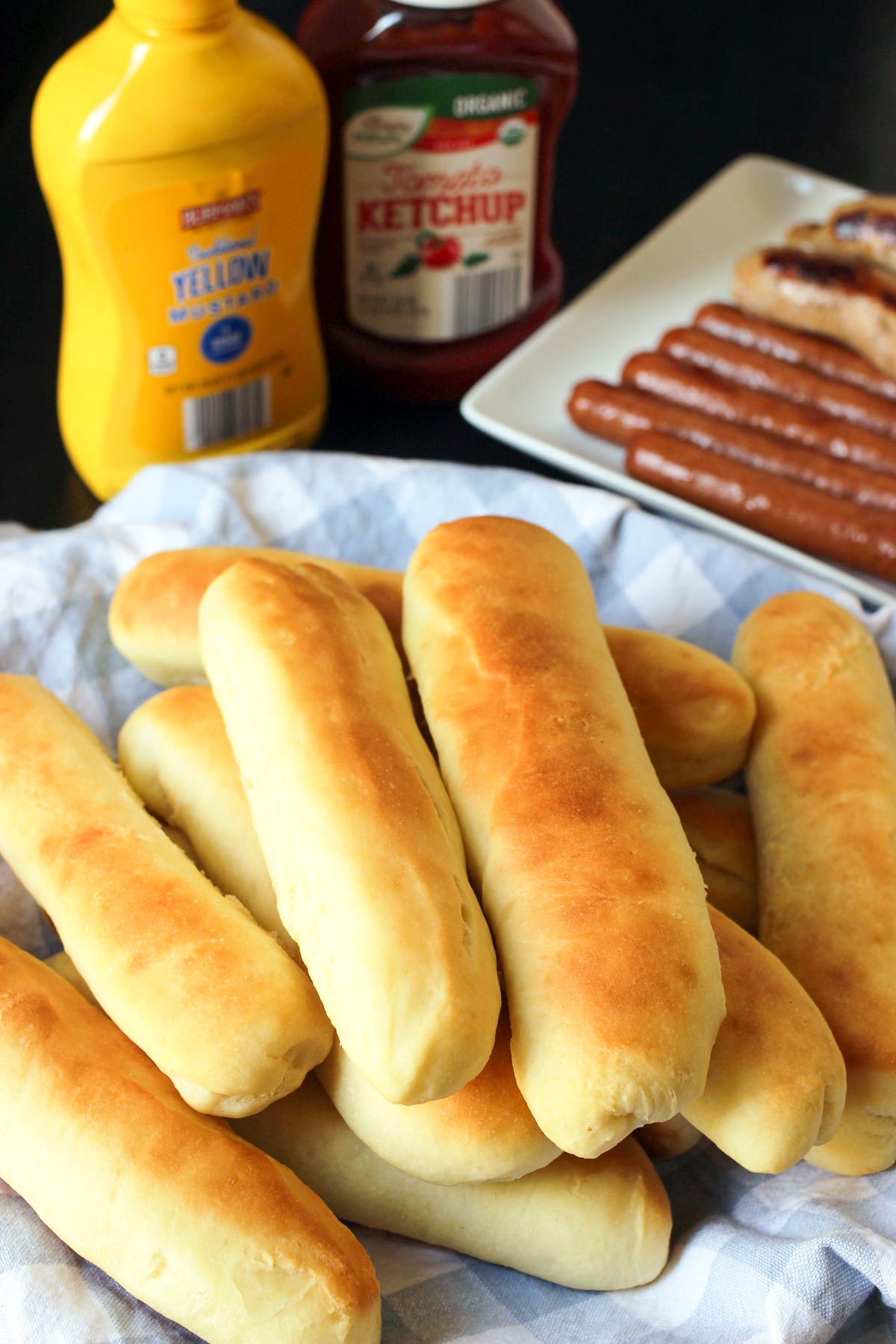 basket of homemade hot dog buns lined with a blue check napkin on a table set with condiments and hot dogs.