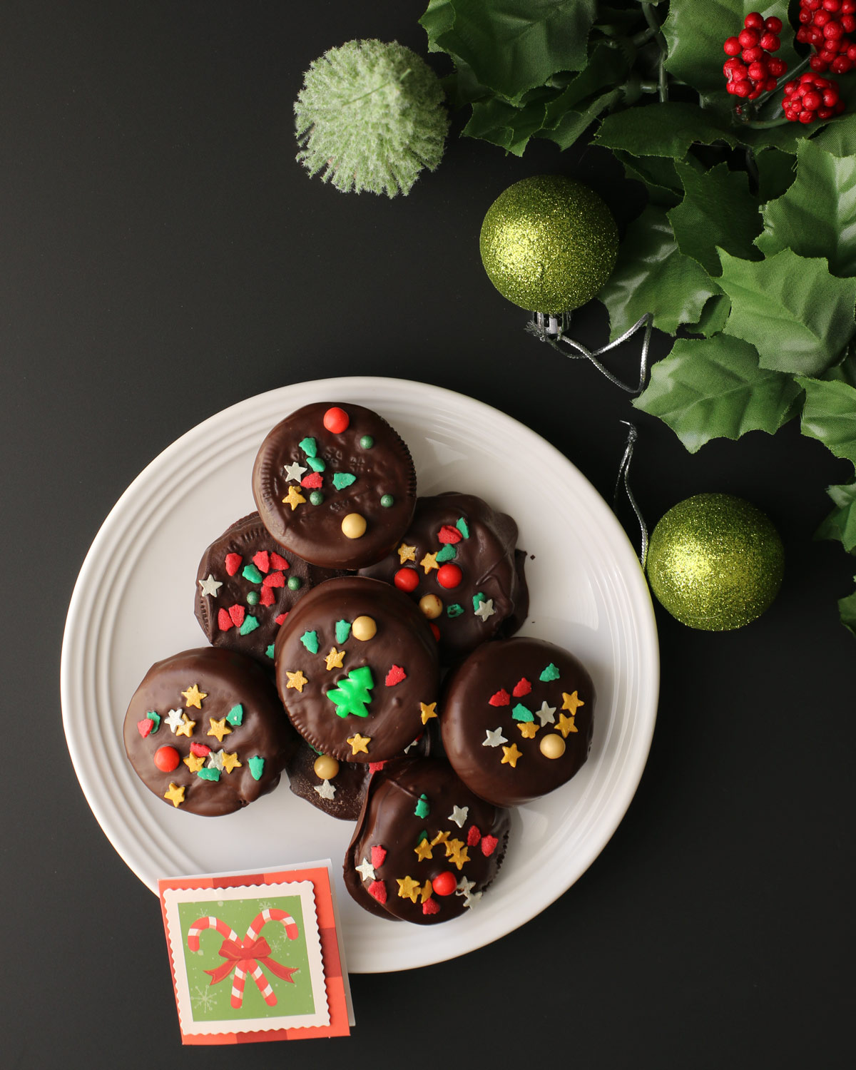plate of chocolate dipped oreos with a christmas tag, ornaments, and holly on table nearby.