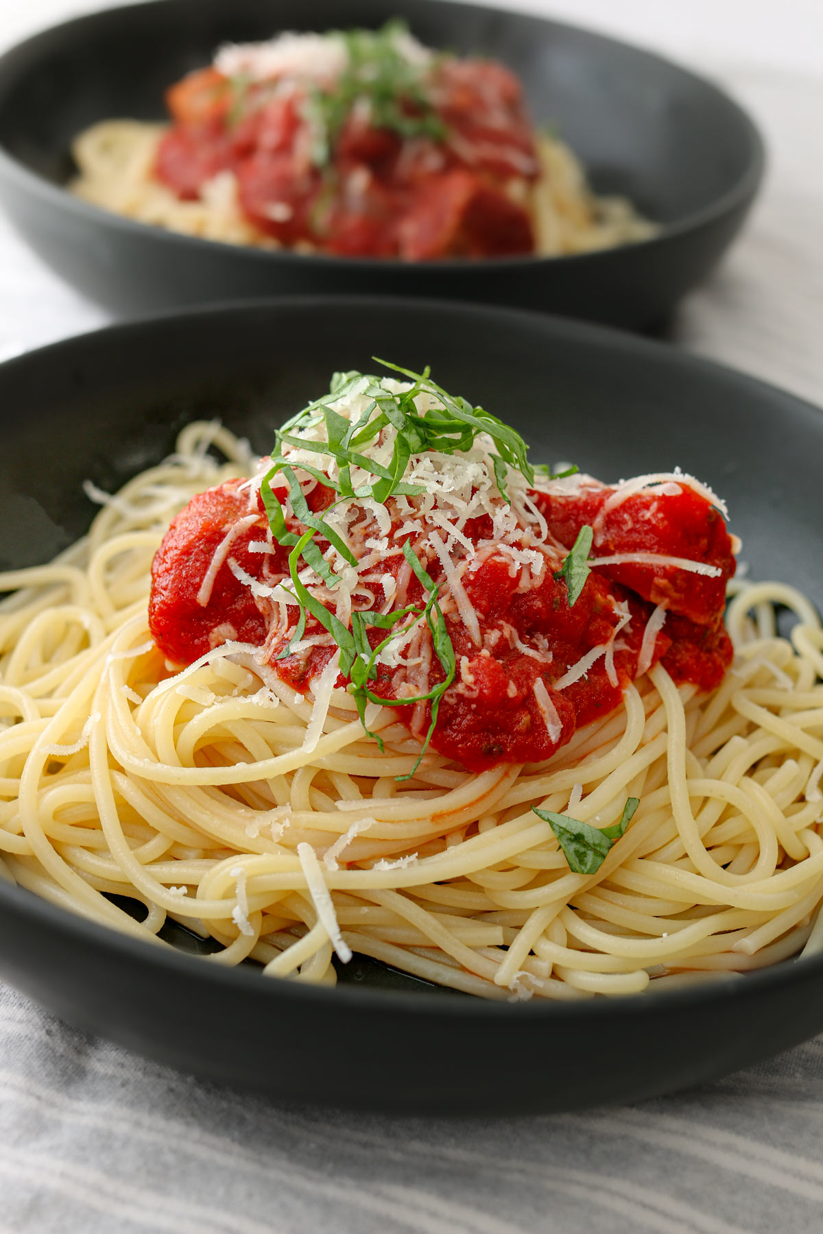 two black pasta bowls with spaghetti topped with sauce, parmesan, and shredded basil.